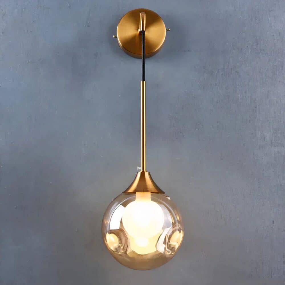 Applique murale suspendue dorée en métal avec boule en verre suspendue. La boule n'est pas lisse mais 2 renfoncements de chaque côté, l'ampoule est allumée, l'applique est fixée sur un mur gris.