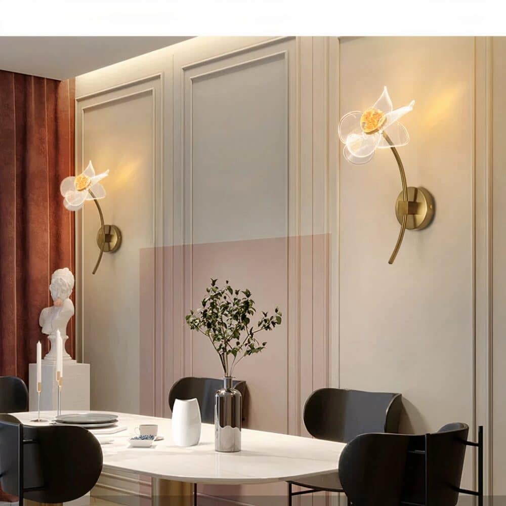 Grande pièce avec table blanche et 4 chaises noires autour. Un vase avec feuillage, bougeoirs, verre blanc posés su la table. A gauche un long rideau marron et une statue blanche devant. Sur le mur en face, 2 appliques murales en forme de fleur avec leur tige sont allumées.