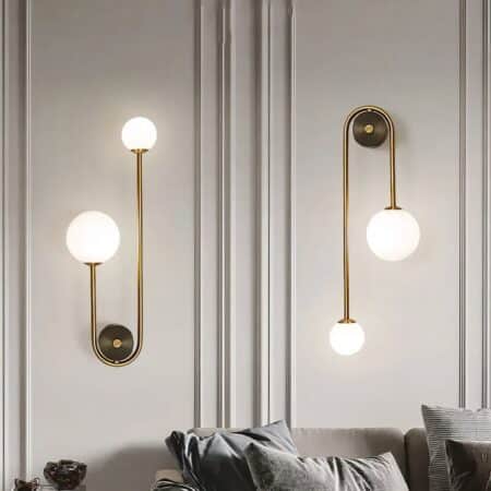 Mur gris où sont fixées 2 appliques murales, support rond doré avec une tige dorée qui passe à gauche et à droite, une plus courte que l'autre. Sur la plus petite, une grosse boule lumineuse, sur la plus longue, une petite boule lumineuse. L'applique de gauche est fixée boules vers le haut, l'applique de droite boules vers le bas. Sous les appliques, on entrevoit un canapé gris avec des coussins assortis.