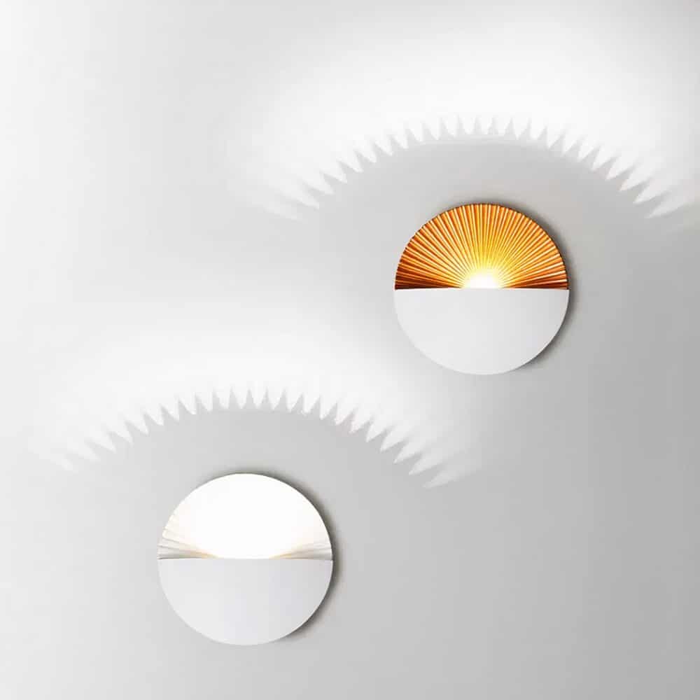 2 appliques murales avec effet coucher de soleil. En haut à droite, l'applique est blanche et la partie intérieure est dorée. En bas à gauche, l'applique est entièrement blanche. Elles sont fixées sur un mur gris et sont allumées et diffusent la lumière sur le mur au-dessus.