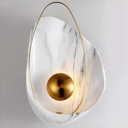 Applique murale en forme de coquillage blanc marbré. A l'intérieur un rond doré donne l'effet d'une perle avec un grand ovale doré autour. Cette applique est fixée sur un mur gris.