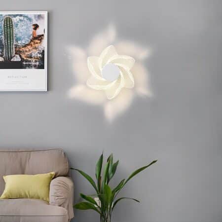 Applique murale en forme de fleur allumée fixée sur un mur gris. A gauche, un tableau et en bas un canapé beige avec un coussin jaune et à sa droite une plante verte.