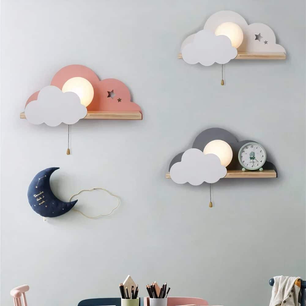 Mur gris avec 3 appliques murales en forme de nuage avec étagère et ampoule allumée visible entre les nuages. Elles sont placées en quinconce, blanche, rose et grise. Sous la rose, il y a une décoration en forme de lune bleu foncé. Sur le bas de l'image on voit des pots à crayons remplis.