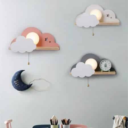 Mur gris avec 3 appliques murales en forme de nuage avec étagère et ampoule allumée visible entre les nuages. Elles sont placées en quinconce, blanche, rose et grise. Sous la rose, il y a une décoration en forme de lune bleu foncé. Sur le bas de l'image on voit des pots à crayons remplis.