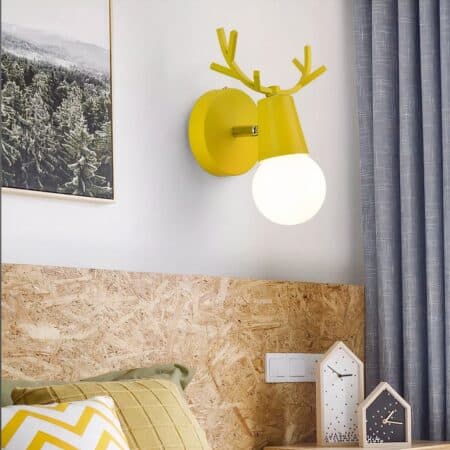 Applique murale jaune design nordique avec des bois de cerf sur le dessus, ampoule allumée, fixée sur un mur blanc entre un tableau sur la gauche et un rideau gris sur la droite. En dessous, un lit beige avec des coussins dans les tons jaunes et beige, des réveils à aiguilles sur la droite.