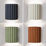 4 appliques murales striées style scandinave, bleu en haut à gauche, blanc en haut à droite, vert en bas à gauche et orange en bas à droite allumée sur mur blanc.