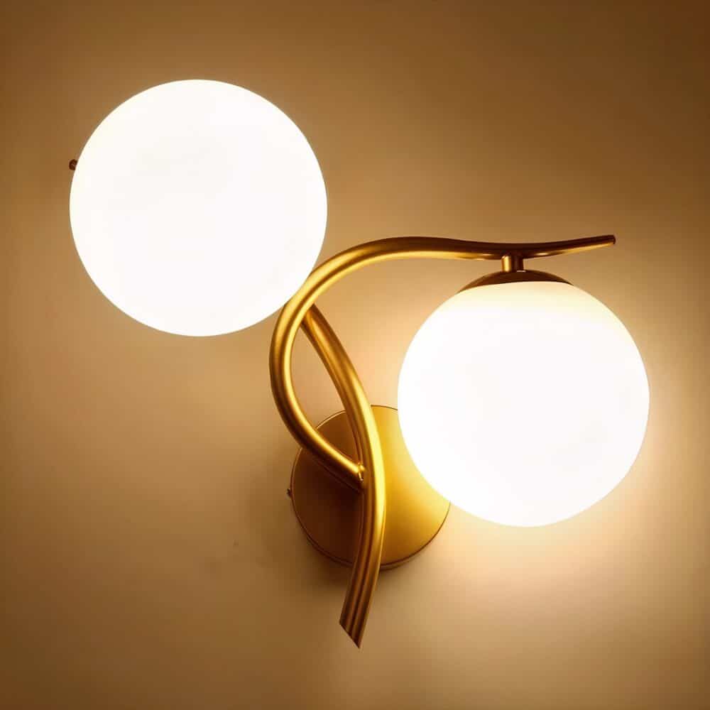 Applique murale en métal doré avec une boule lumineuse au-dessus de la tige verticale et l'autre boule sous la tige horizontale côté droit. Les 2 boules lumineuses sont allumées. Fixée sur un mur beige.