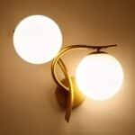 Applique murale en métal doré avec une boule lumineuse au-dessus de la tige verticale et l'autre boule sous la tige horizontale côté droit. Les 2 boules lumineuses sont allumées. Fixée sur un mur beige.