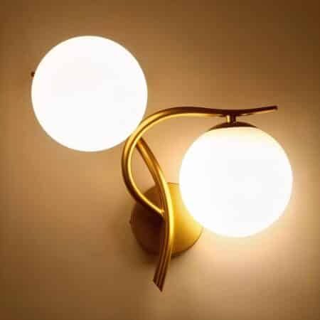 Applique murale en métal doré avec une boule lumineuse au-dessus de la tige verticale et l'autre boule sous la tige horizontale côté droit. Les 2 boules lumineuses sont allumées. Fixée sur un mur beige.