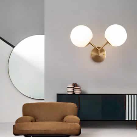 Applique murale dorée avec 2 boules lumineuses tenue par une tige dorée en forme de V. Fixée sur un mur gris au-dessus d'un meuble bas noir Un petit fauteuil marron est devant. A gauche au fond un grand miroir rond.