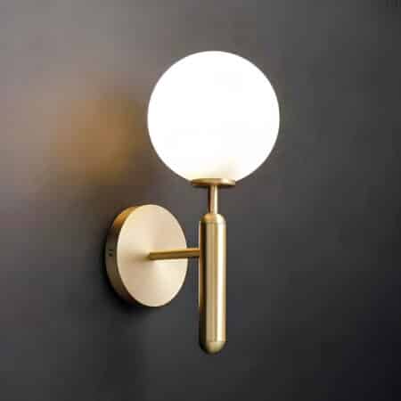 Applique murale dorée avec support rond au mur et tige dorée qui tient une boule lumineuse. Fixée sur un mur noir.