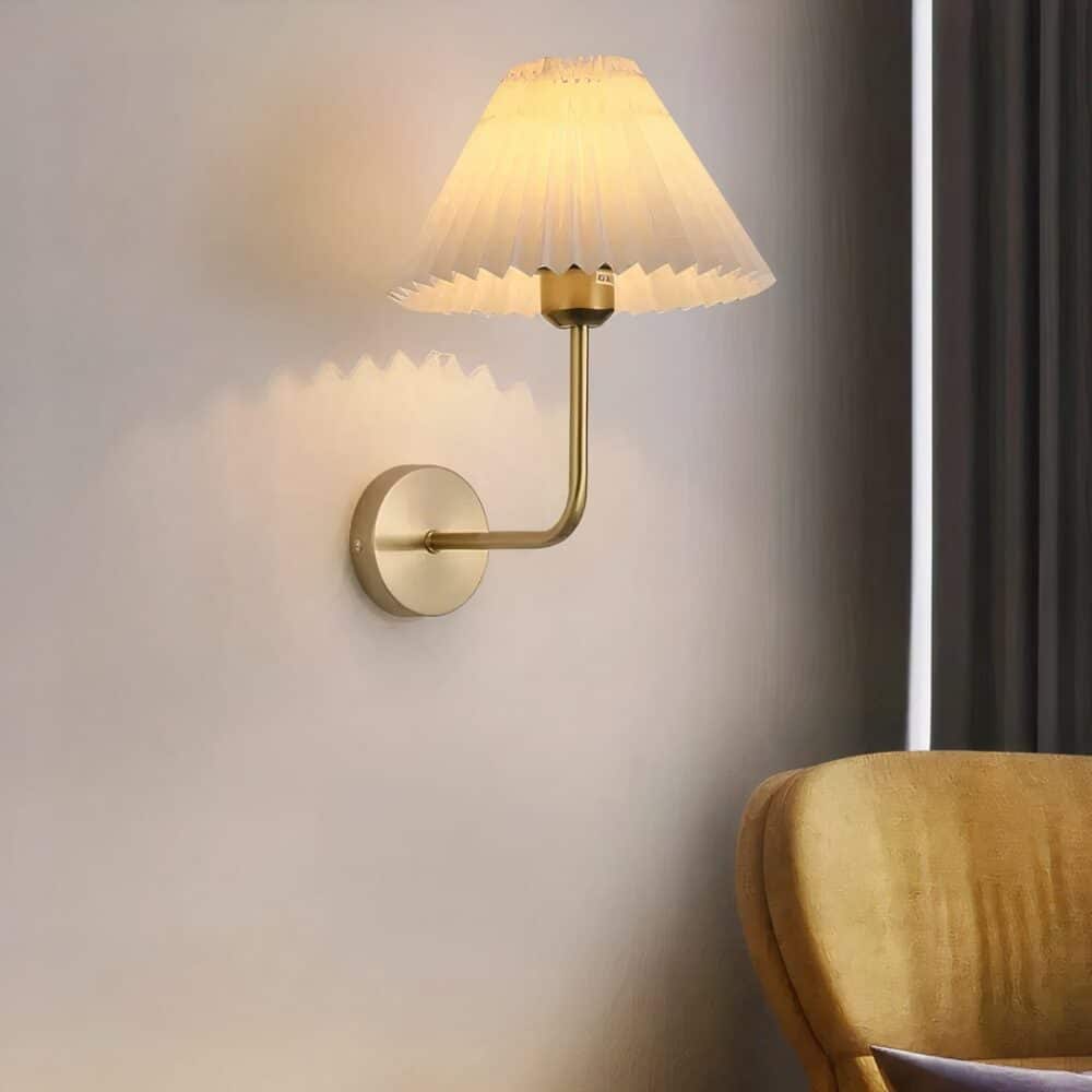 Applique murale de salon vintage avec support rond et bras coudé doré tenant un abat-jour blanc en tissu plié en forme d'accordéon. Lumière chaude diffusée. Fixée sur un mur gris, un fauteuil jaune moutarde à droite et un rideau gris foncé derrière.