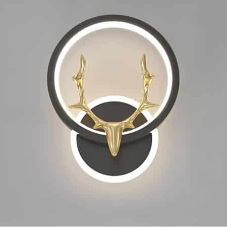 applique murale noire, le support est rond avec une cercle de lumière autour. Il tient un cercle lumineux dans un cercle noir sur lequel est posé une tête de cerf doré. Sur fond gris.