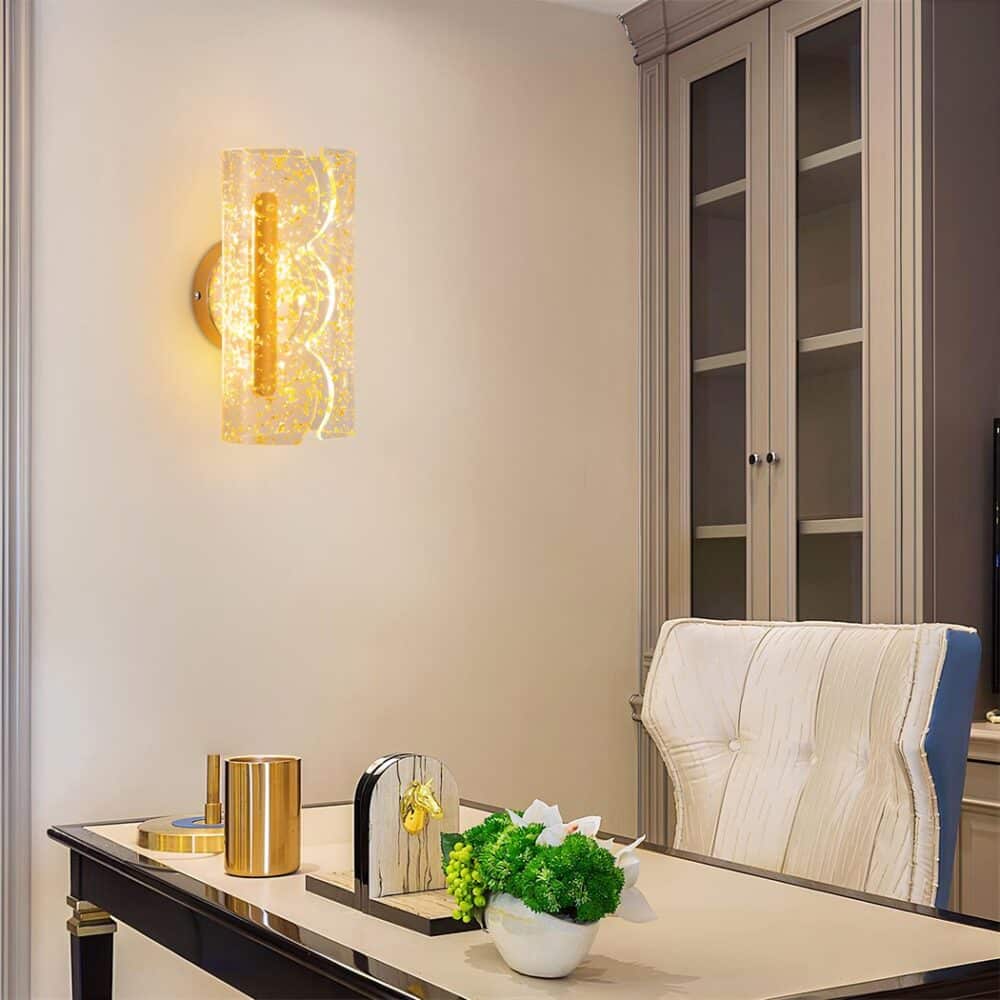 Applique murale de forme rectangulaire arrondi ouvert sur le devant en forme de 3 ponts dans la verticale. Le support est en métal doré, le reste est en acrylique transparent avec des incrustations imitant des feuilles d'or. La lumière se diffuse à travers l'acrylique. Fixée sur un mur face à un bureau avec les pieds et l'encadrement noir, le plateau est beige. Dessus, il y a une petite plante verte, un cale livre, un pot à crayons doré. Un fauteuil en simili cuir beige et gris et derrière une fenêtre avec encadrement gris.