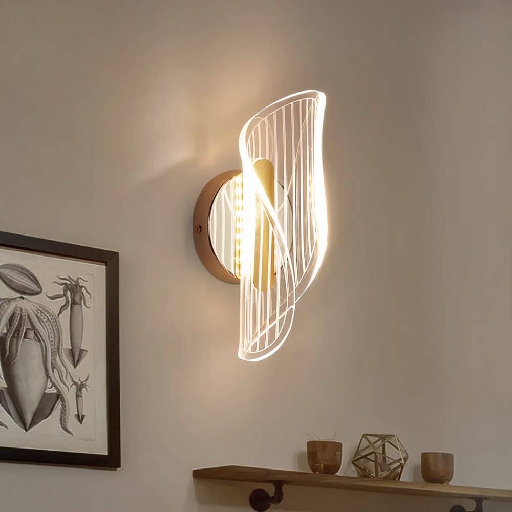 Applique murale en acrylique ondulée sur un support rond doré. La lumière est diffusée à travers l'acrylique. L'applique murale est fixée sur un mur beige, au-dessus d'une étagère en bois avec 3 petits pots. A gauche de l'applique murale, il y a un cadre blanc et noir.