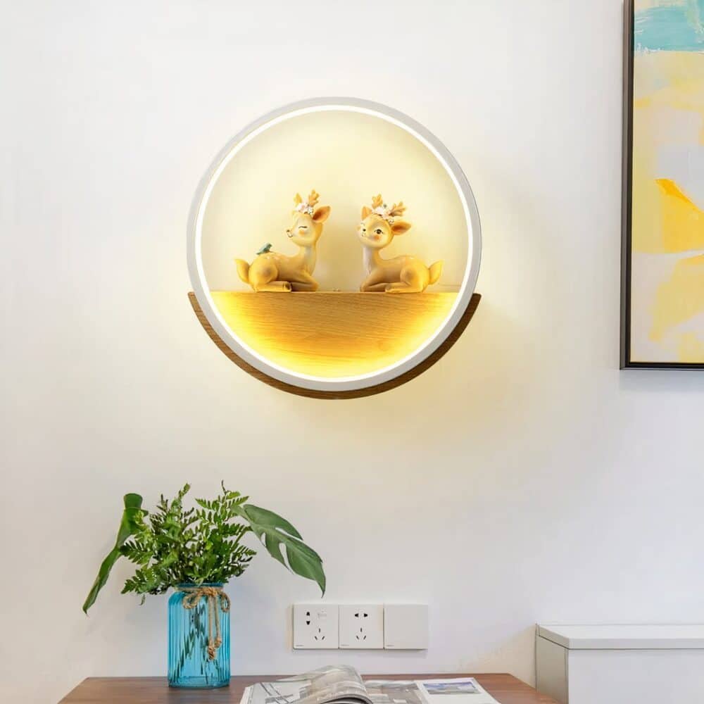 Applique murale en forme de cercle blanc. A l'intérieur, il y a un support en demi lune en bois avec 2 cerfs posés face à face. L'applique murale est allumée et fixée sur un mur blanc, un cadre coloré à droite et en dessous un meuble marron avec un vase bleu et des feuillages dedans et un magazine.
