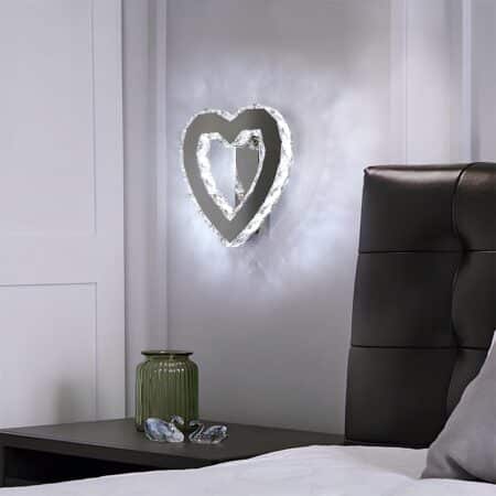 Applique murale en forme de cœur creux de couleur argenté avec l'intérieur et l'extérieur des contours lumineux imitant des diamants. La lumière est froide. Fixée au-dessus d'une table de chevet avec un pot vert. A droite, une tête de lit simili cuir noir avec literie blanche.