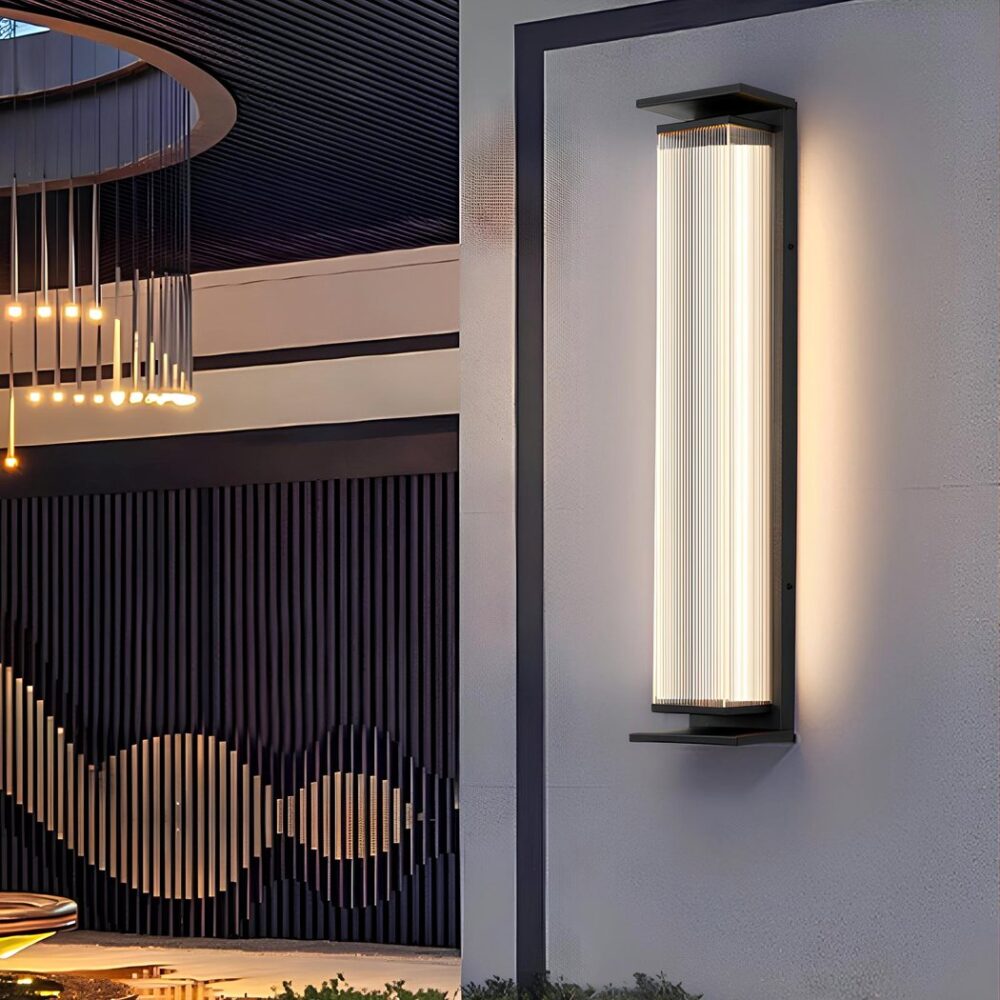 Applique murale rectangulaire avec structure en acier noir et capot lumineux en plastique laissant passer la lumière au travers. Fixée sur un mur blanc avec liseré noir. Sur la gauche, des suspensions lumineuses en tiges tenues par un plafond ajouré.