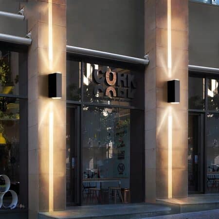 Devanture de magasin avec des vitrines encadrées par des piliers en béton où sont fixées une applique murale sur chacun. L'applique murale est rectangulaire de couleur noire et diffuse un faisceau au-dessus et en-dessous en lumière chaude.