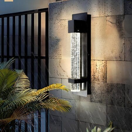 Applique murale extérieure en forme de tube à bulles dans une structure en aluminium noir fixé sur un mur en pierre. A gauche un grand portail noir, une partie de palmier devant le mur et la pluie qui tombe.