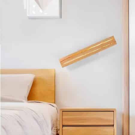 Applique murale moderne rectangulaire en bois avec stries qui laissent passer la lumière sur l'avant. Elle est penchée vers la gauche où se trouve un lit avec tête de lit marron et une literie blanche, une table de chevet en bois sous l'applique murale et un cadre blanc au-dessus du lit.