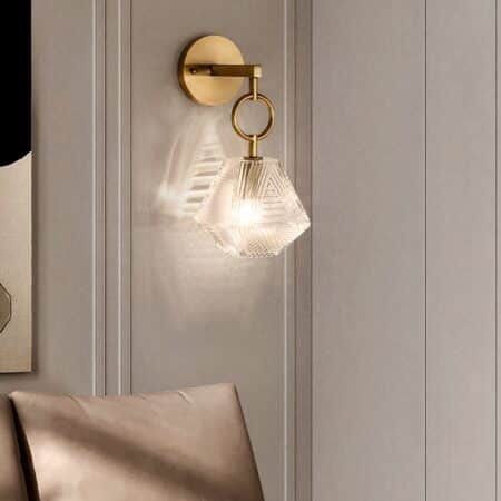 Applique murale dorée avec polygone en verre suspendu avec ampoule allumée. Fixée sur un mur gris au-dessus d'un fauteuil marron.