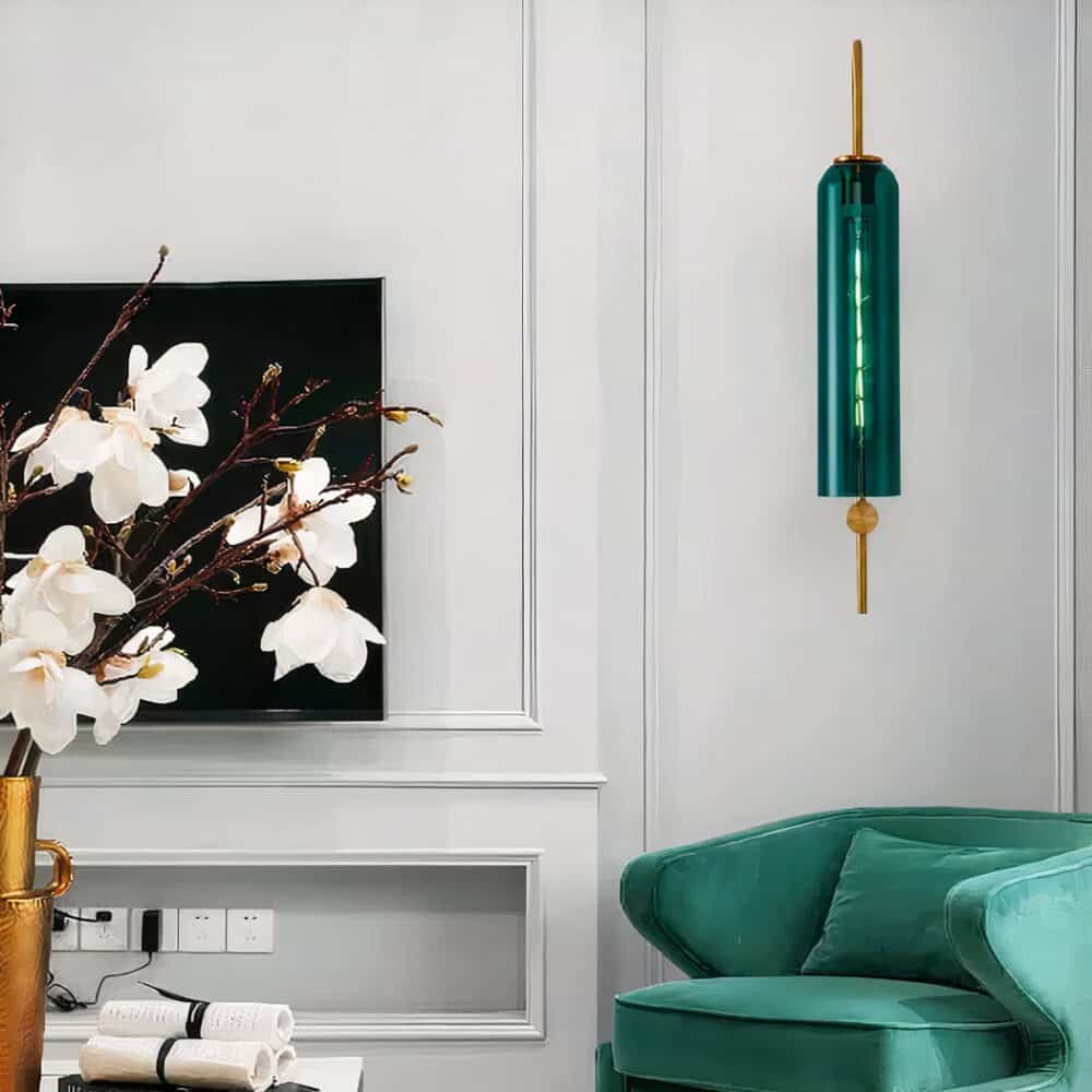 Applique murale tube en verre style art déco allumée et accrochée sur un mur blanc de salon, au-dessus d'un fauteuil vert forêt.