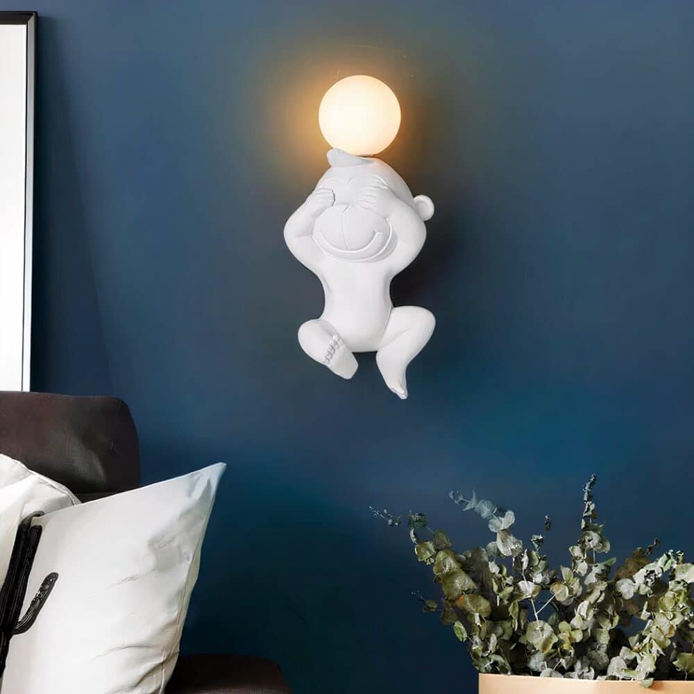 Applique murale blanche en forme de singe avec les mains sur les yeux, une ampoule allumée sur la tête. Fixée sur un mur bleu canard au-dessus d'une plante verte.
