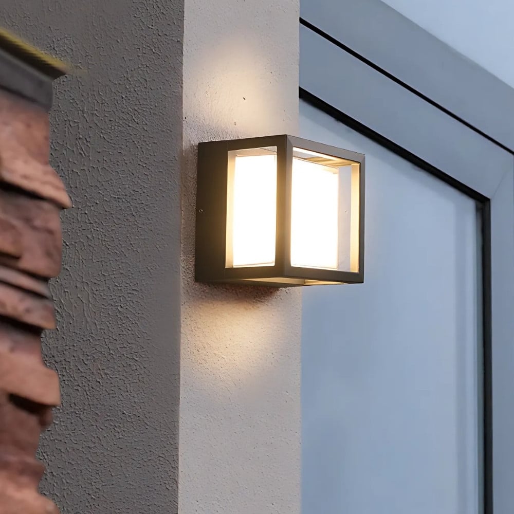 Applique luminaire extérieur en forme de cube en métal noir et au centre un cube en crylique diffusant la lumière. Fixée sur un mur blanc en extérieur avec une vitre sur la droite.