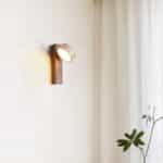 Applique murale style Japandi marron, avec lumière orientable. Accrochée à un mur clair, dans une pièce avec des rideaux et une plante.